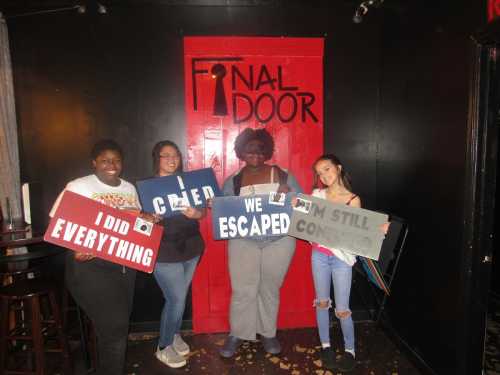 Four people hold signs in front of a red door labeled "FINAL DOOR," celebrating their escape from an escape room.