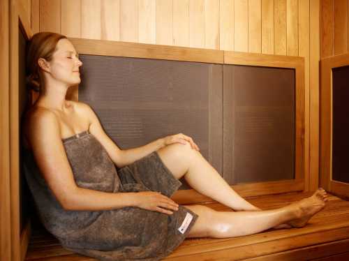 A woman relaxes in a wooden sauna, sitting against the wall with a towel wrapped around her.