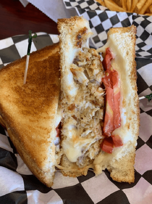 A close-up of a grilled sandwich with melted cheese, shredded meat, and tomato slices, served on a checkered paper.