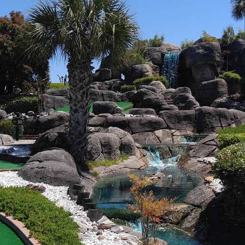 A scenic mini-golf course featuring palm trees, rocky landscapes, and a small waterfall surrounded by lush greenery.