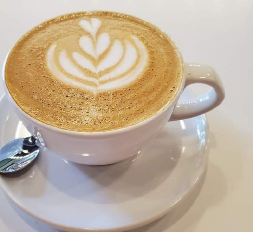 A cup of latte with intricate leaf latte art, placed on a white saucer with a small spoon beside it.