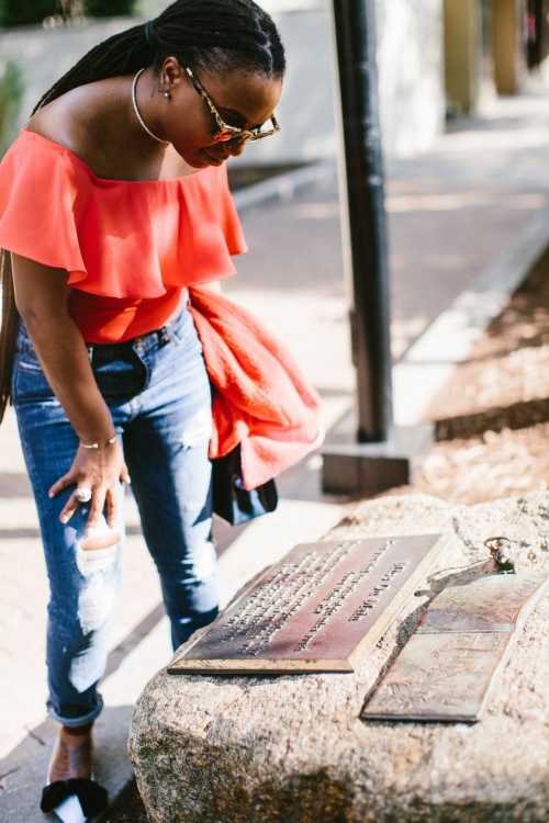 A woman in a red off-shoulder top examines engraved plaques on a rock in an outdoor setting.