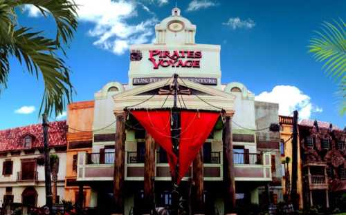 Exterior of Pirates Voyage, featuring a large ship with a red sail in front of a themed building against a blue sky.