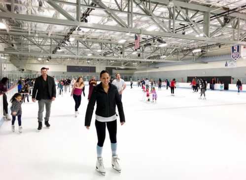 A woman in skates smiles on an ice rink, surrounded by skaters of all ages enjoying the activity.