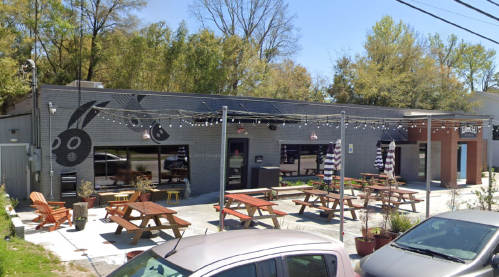 A modern restaurant exterior with picnic tables, string lights, and a mural of playful rabbits on the gray wall.