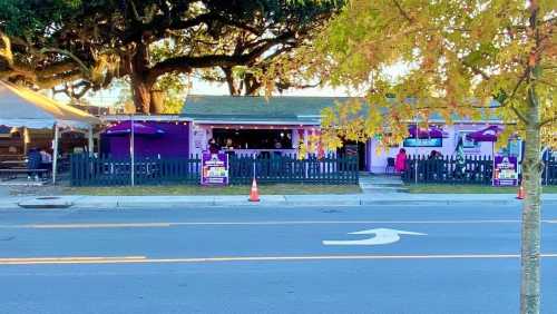 A colorful restaurant with purple accents, outdoor seating, and a fenced area, set along a tree-lined street.