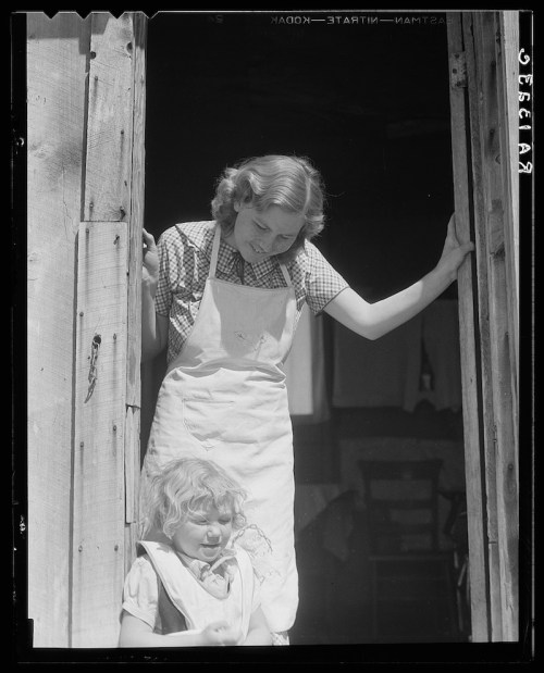A woman in an apron leans in a doorway, watching a young girl playing outside.