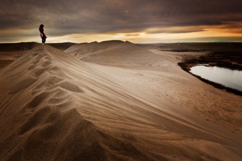 A person stands on a sand dune at sunset, overlooking a river and a vast landscape under a cloudy sky.