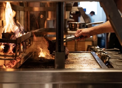 A chef uses tongs to handle meat over an open flame in a busy kitchen. Flames rise from the grill.