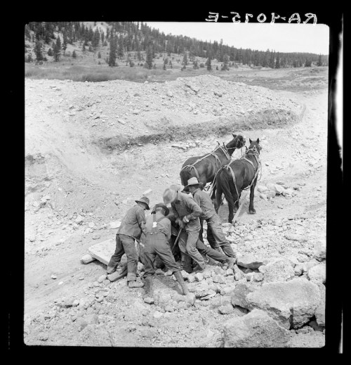 A group of workers in hats is gathered around a large object on a rocky terrain, with two horses nearby.