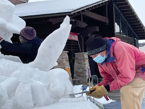 Two artists sculpt ice outside a building, one carving with a tool while the other works in the background.