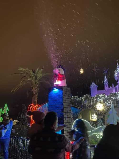 A festive scene with colorful lights, a snowman statue, and people enjoying a holiday display under a night sky.