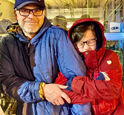 A man and a woman embrace warmly, both wearing colorful jackets, smiling in a cozy indoor setting.