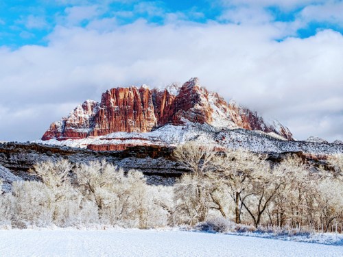 Snow-covered landscape with a majestic red rock formation under a partly cloudy blue sky.