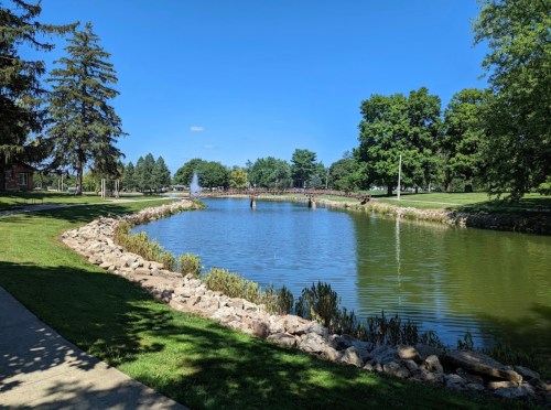 A serene park scene featuring a pond, lush greenery, and a bridge in the background under a clear blue sky.