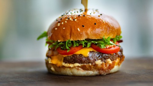 A close-up of a juicy burger with lettuce, tomato, cheese, and a sesame seed bun, garnished with a toothpick.