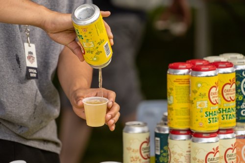 A person pours a yellow beverage from a can into a small cup, with colorful cans stacked in the background.