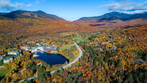 Aerial view of a vibrant autumn landscape with colorful trees, a lake, and mountains in the background.