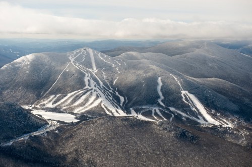 Aerial view of snow-covered mountains with ski trails and a frozen lake below, under a cloudy sky.