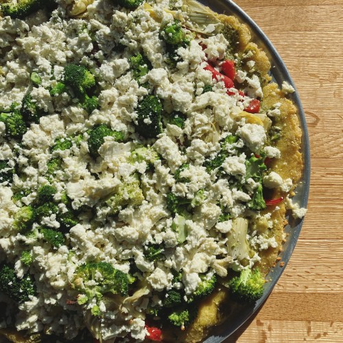 A close-up of a colorful vegetable dish topped with crumbled cheese, featuring broccoli, red peppers, and a golden crust.