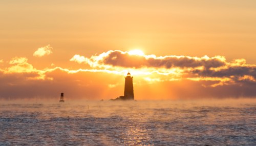 A lighthouse silhouetted against a vibrant sunset, with mist rising from the water.