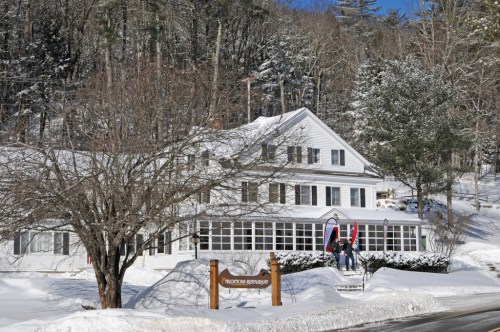 A snowy scene featuring a white house with a sign reading "Restaurant" and trees in the background.