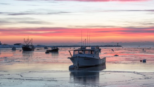 A serene sunset over icy waters, featuring fishing boats silhouetted against vibrant pink and orange skies.