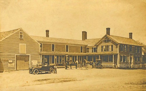 Historic sepia-toned image of a two-story building with horse-drawn carriages and an early automobile parked outside.