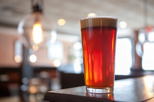 A glass of amber beer with a frothy head, set on a wooden surface in a softly lit bar.