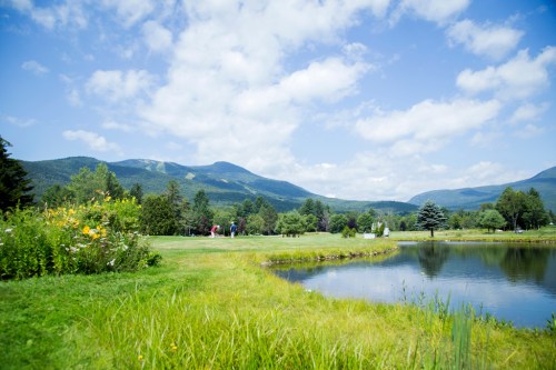 A scenic view of a golf course with mountains in the background, a pond, and lush greenery under a blue sky.