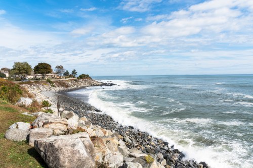A rocky shoreline with waves crashing, under a blue sky with scattered clouds and a distant coastal landscape.