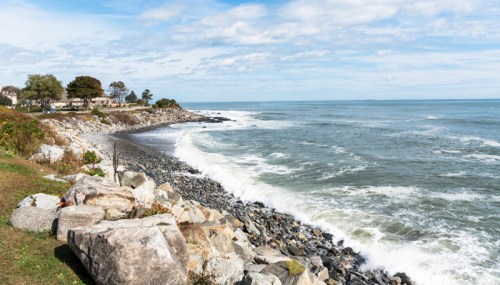 A rocky shoreline with waves crashing, under a blue sky with scattered clouds and a few trees in the background.