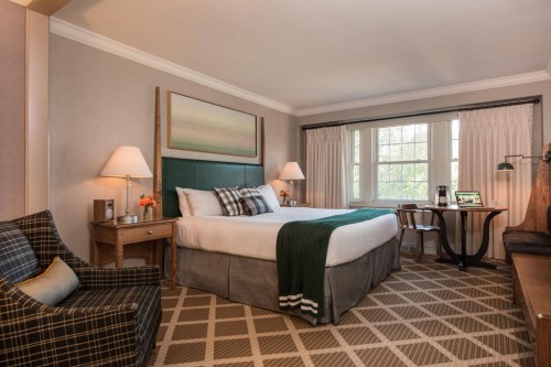 Cozy hotel room featuring a king bed, armchair, desk, and large window with natural light. Warm, inviting decor.