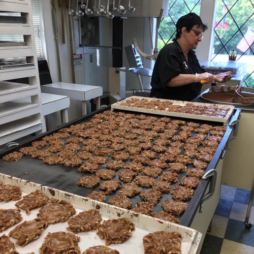 A woman prepares chocolate-covered treats in a kitchen filled with trays of confections.
