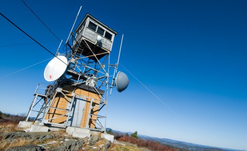 A tall communication tower with antennas, set against a clear blue sky and distant mountains.