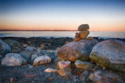 A stack of smooth stones on larger rocks by the shore, with a calm sea and sunset in the background.