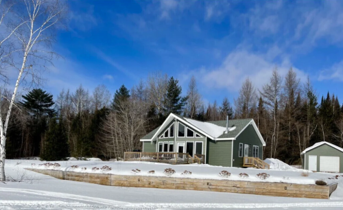 A modern green house with a large deck, surrounded by snow and trees under a blue sky.