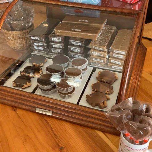 A wooden display case filled with various chocolates, including turtle candies and fudge, on a wooden table.