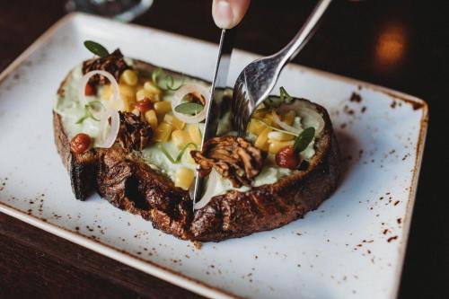 A hand using a fork and knife to cut into a slice of toast topped with avocado, corn, and garnishes on a white plate.