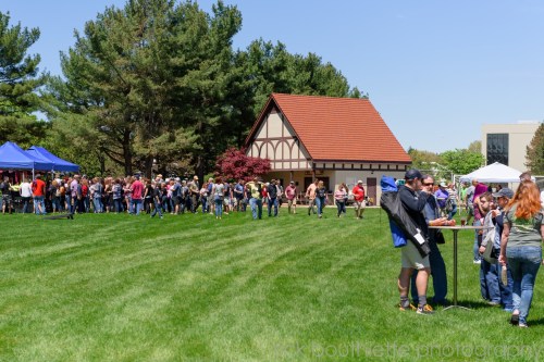 A large crowd gathers on a grassy area near a building, enjoying an outdoor event with tents and tables.