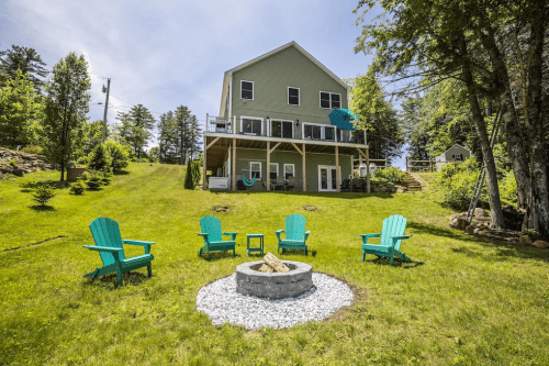 A green house with a deck overlooks a grassy yard featuring a fire pit surrounded by teal chairs. Trees in the background.