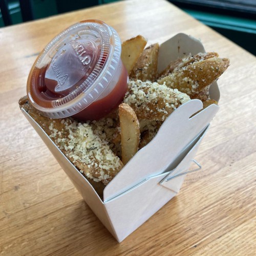A takeout box filled with seasoned potato wedges and a small cup of ketchup on top.