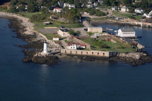Aerial view of a coastal area featuring a lighthouse, buildings, and rocky shoreline near a calm body of water.