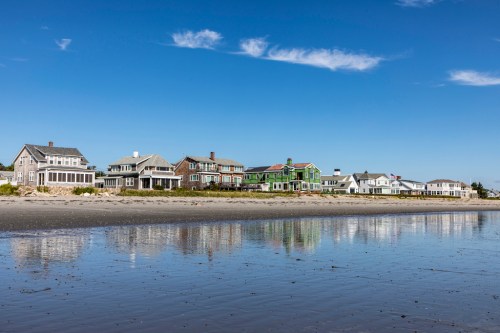 A row of beach houses along a sandy shore, reflecting in the calm water under a clear blue sky.