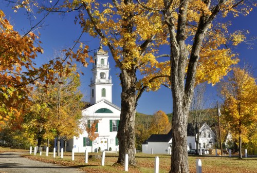 A white church with a tall steeple surrounded by vibrant autumn trees and a clear blue sky.