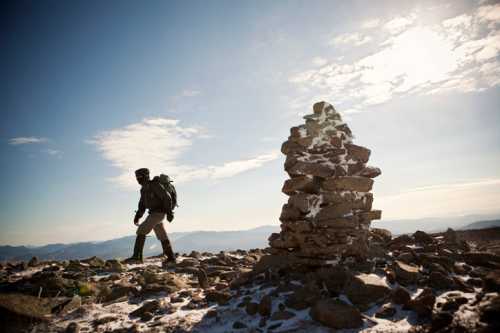 A hiker with a backpack walks past a stone cairn on a rocky mountain summit under a clear blue sky.