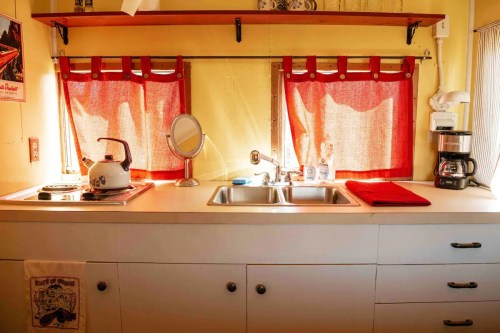 A cozy kitchen with yellow walls, red curtains, a kettle, coffee maker, and a double sink. Bright and inviting atmosphere.