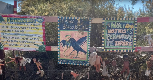 Colorful signs on a fence with welcoming messages and artwork, surrounded by dolls and greenery.