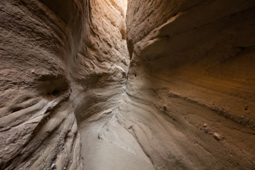 Narrow canyon with smooth, layered rock walls and soft sand at the base, illuminated by natural light from above.