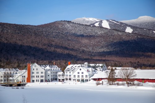 A snowy landscape featuring a large white hotel near a frozen lake, with mountains in the background under a clear blue sky.